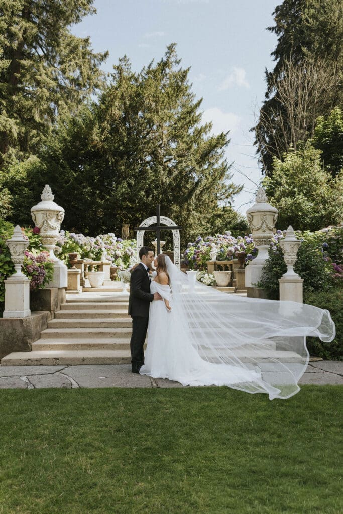 Bride and groom standing in the English Sunken Garden location for the wedding ceremony with veil fluttering in the wind, photographed by Seattle wedding photographer Valen Lee Photography.