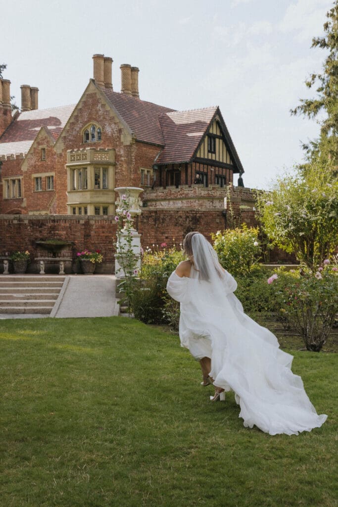 Bride walking through the gardens toward a historic brick estate in her flowing wedding gown and veil.